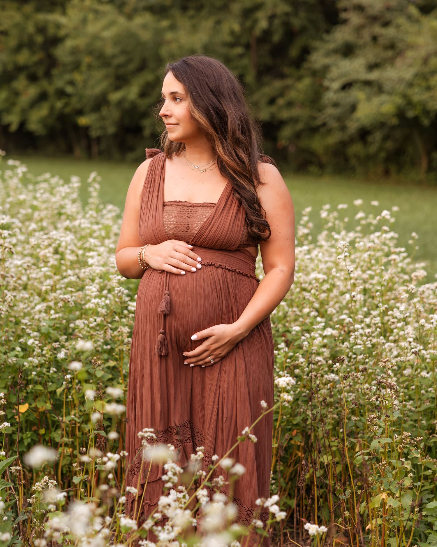 Pregnant woman in rust brown maxi dress standing in wildflower field during outdoor maternity photo session.