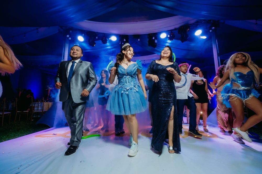 Guests dancing joyfully at a quinceañera celebration under blue stage lights in an elegant tent venue.