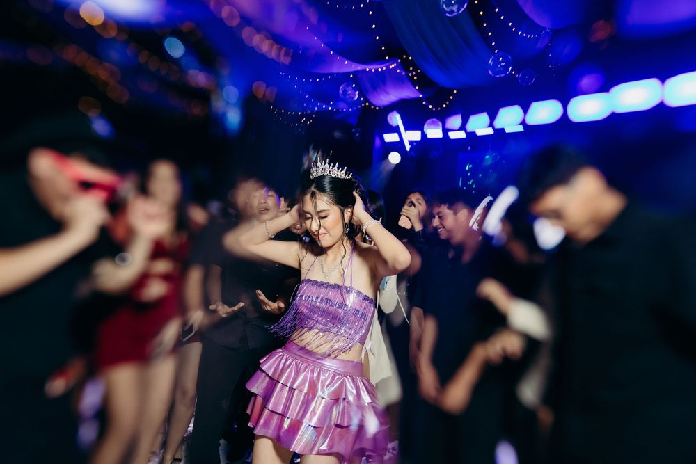 Young woman in purple dress and tiara dancing at a vibrant nightclub party with colorful lights.