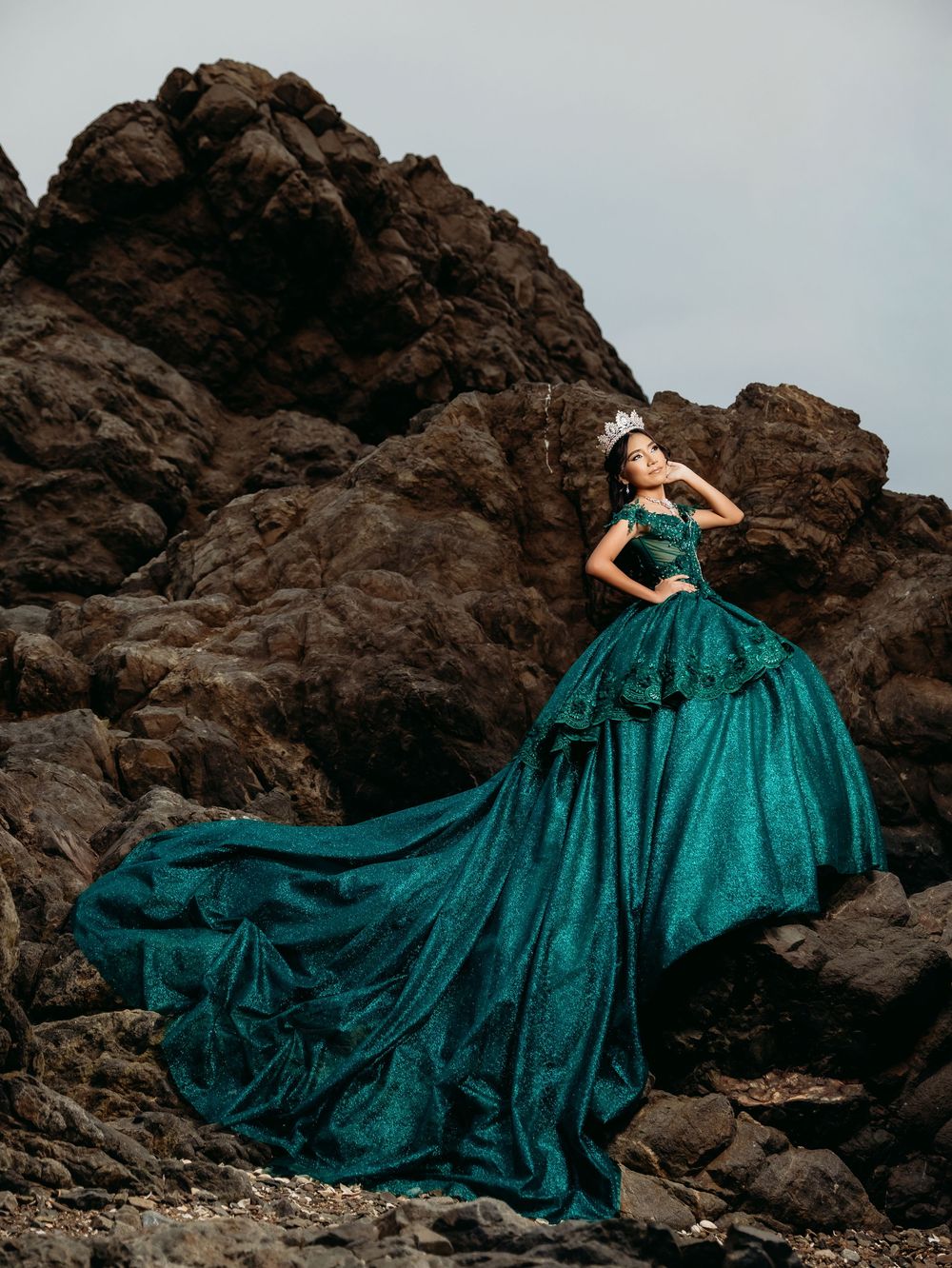 Woman in emerald green ball gown with crown poses dramatically on rocky coastal cliffs.