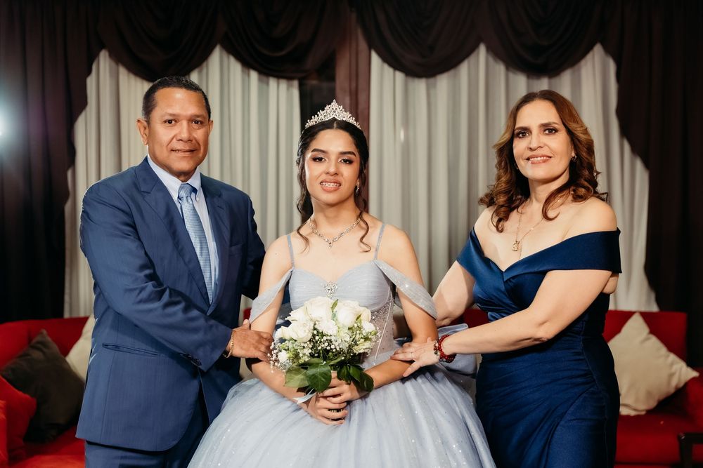 A young woman in a blue quinceañera gown with tiara holds white roses, flanked by her parents in formal attire.