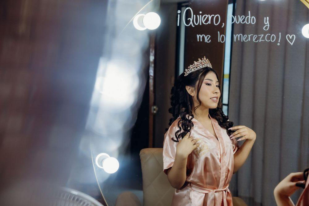 Young woman in pink satin robe and tiara poses before a mirror with Spanish motivational quote during quinceañera preparation.