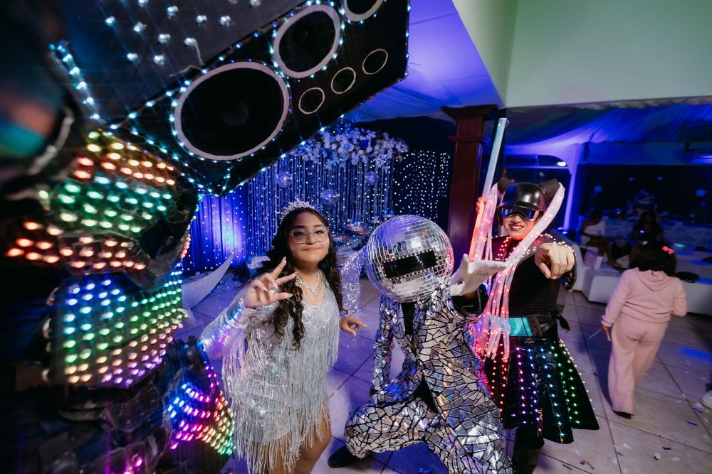 Woman in silver outfit poses with disco ball costume character at colorful neon-lit party event.
