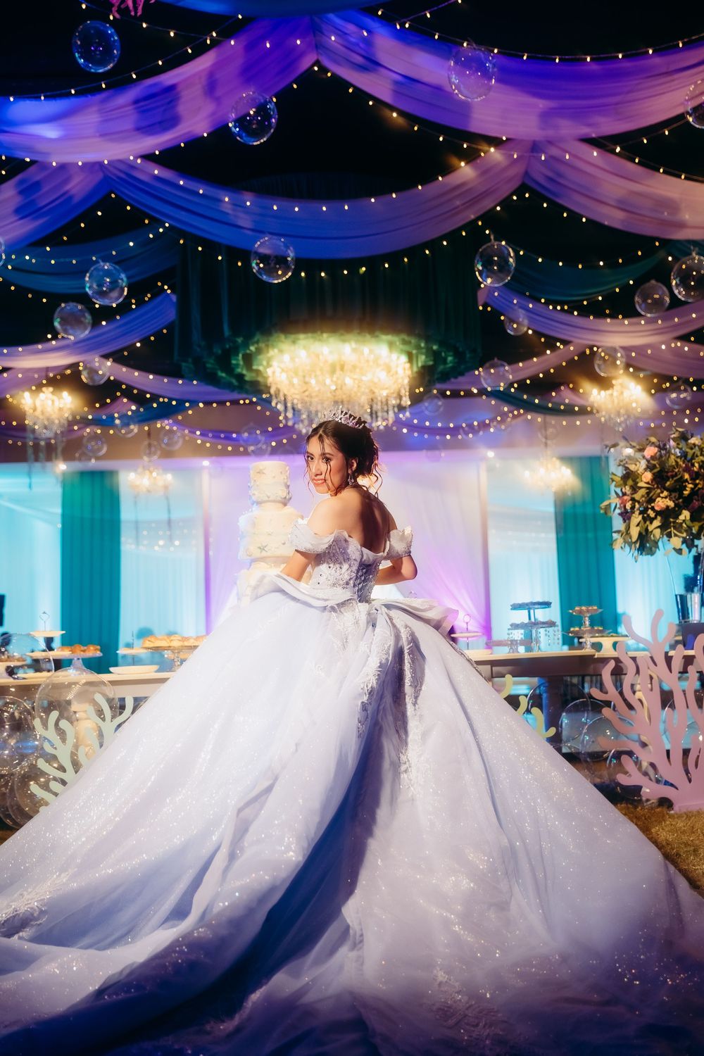 Young woman in elegant white ball gown poses at a beautifully decorated quinceañera venue with bubbles and fairy lights.