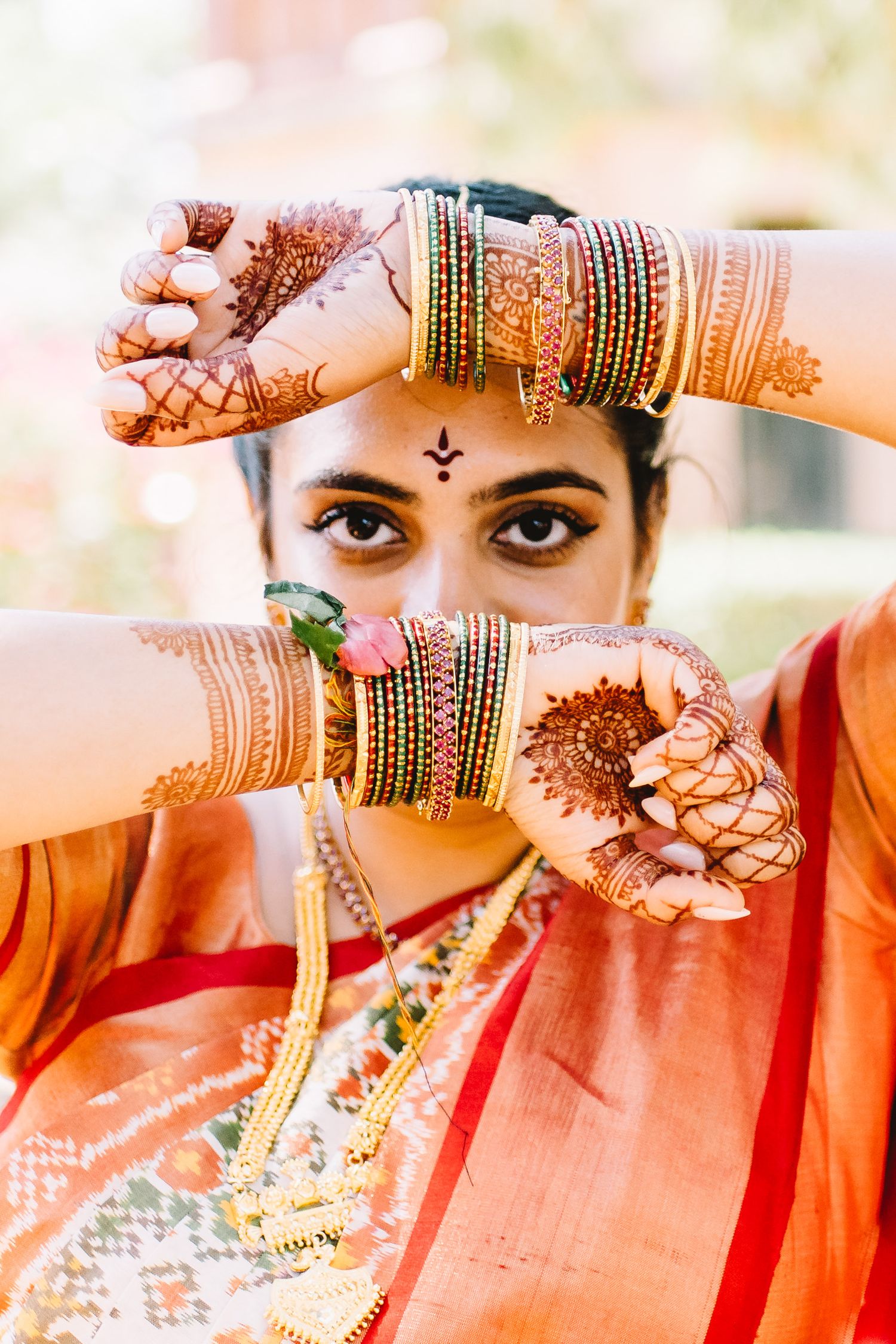 Bride in a red and orange saree framing her eyes with henna-covered hands, wearing stacked bangles and gold necklaces.