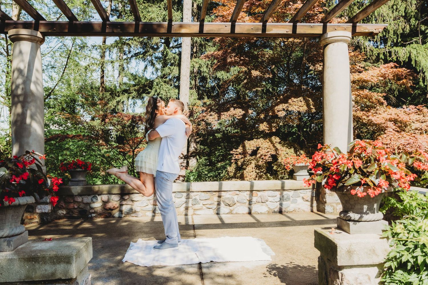 Couple embracing as he lifts her in a romantic engagement photo on a garden patio with stone pillars, red flowers and autumn foliage.