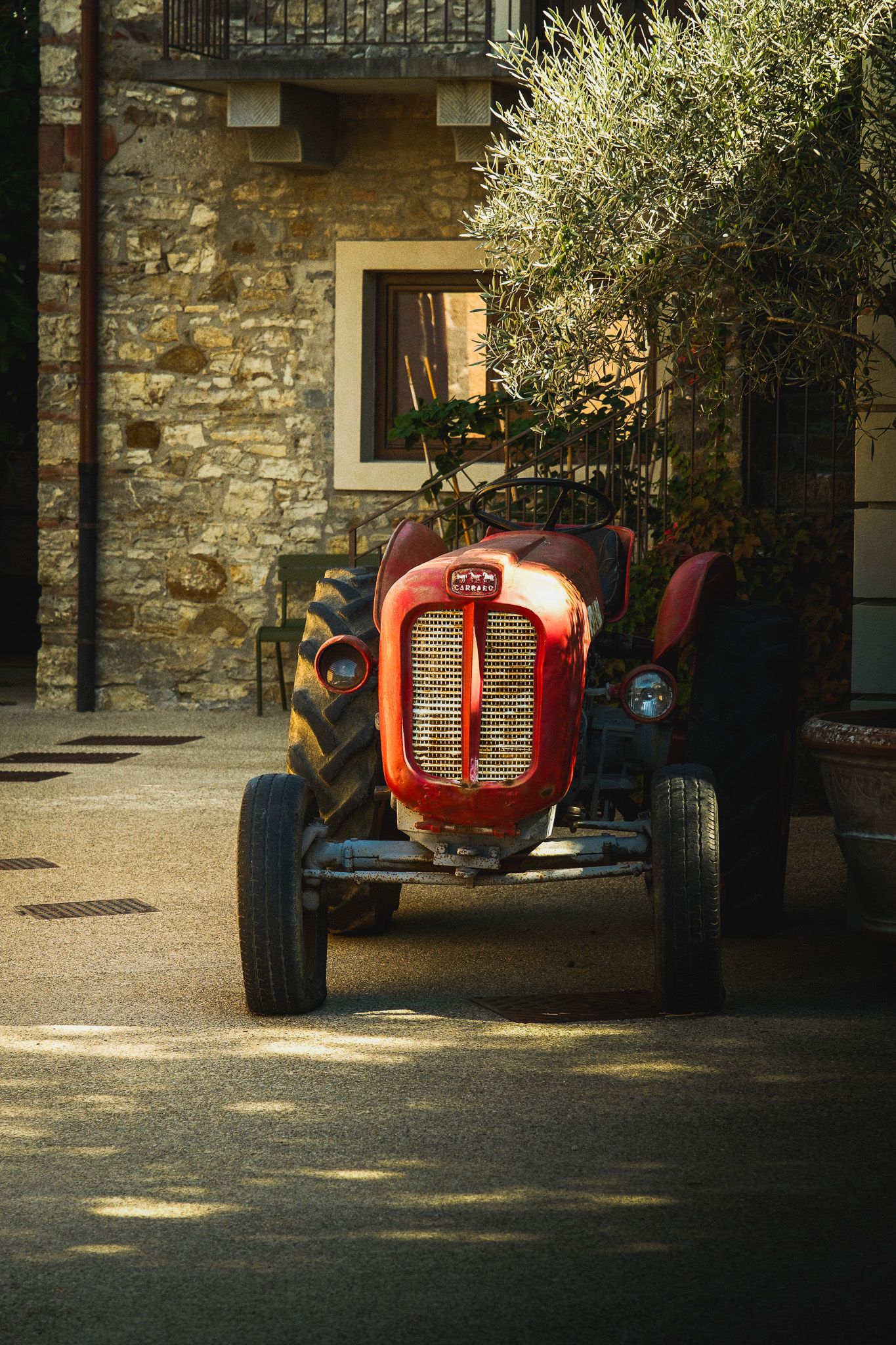 Vintage red tractor parked in a sunlit courtyard beside a rustic stone building and an olive tree.