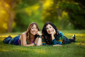 Two smiling women lying on green grass in a sunny park, posing together for a portrait photo.