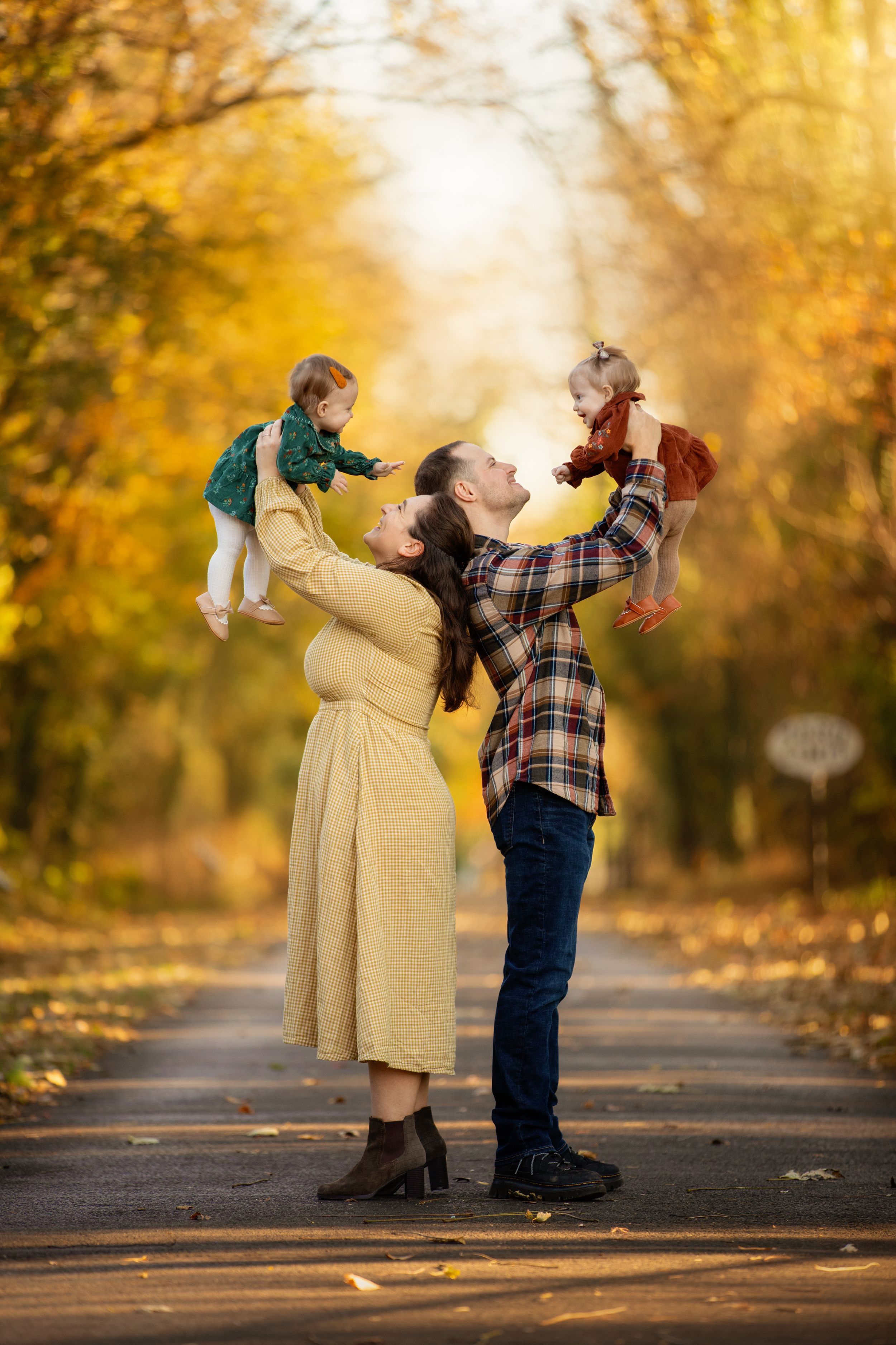 A playful autumn scene in a park with golden leaves as two people hold up teddy bears on a tree-lined path.
