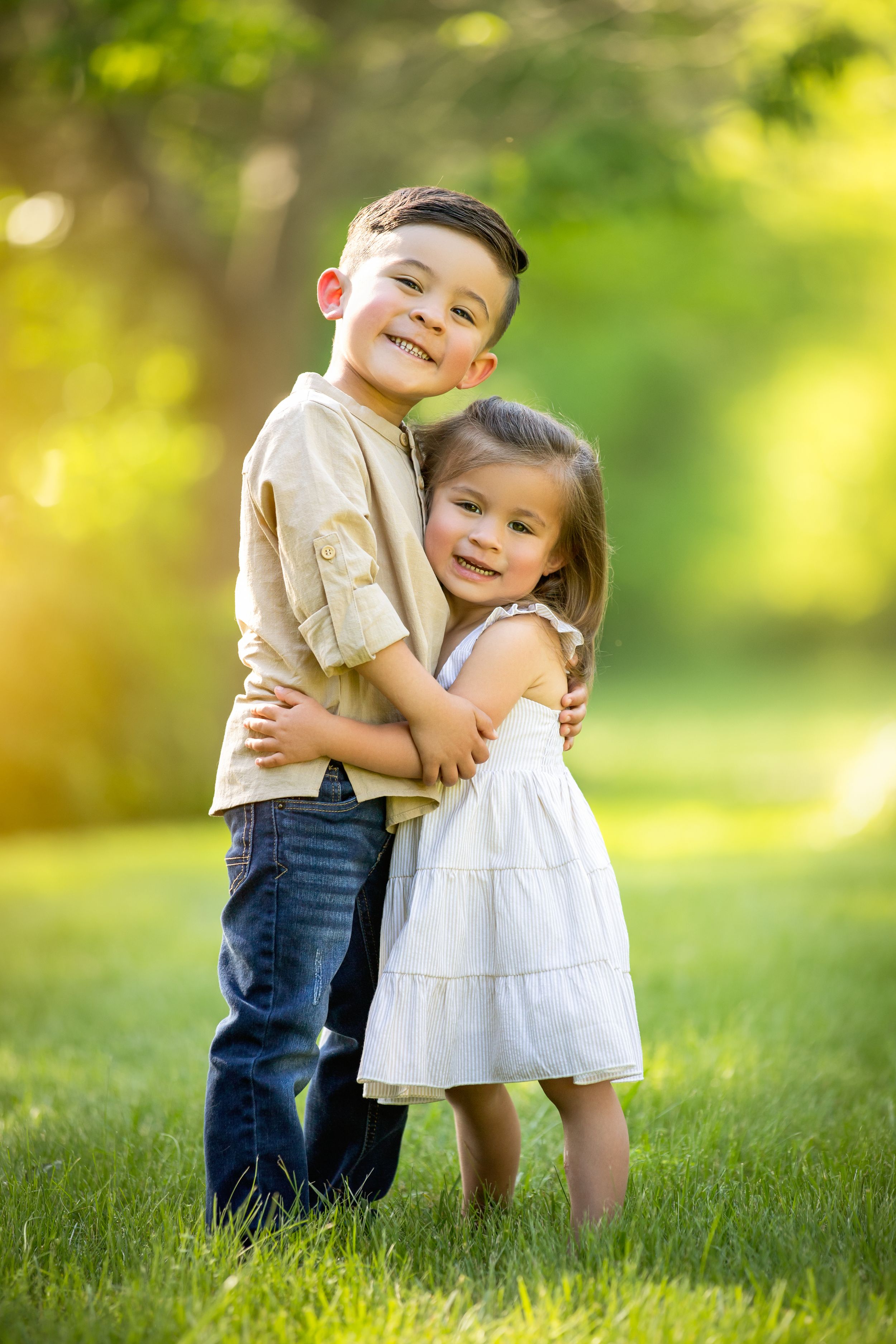 Two young children hugging and smiling on grass with golden sunlit bokeh in background.