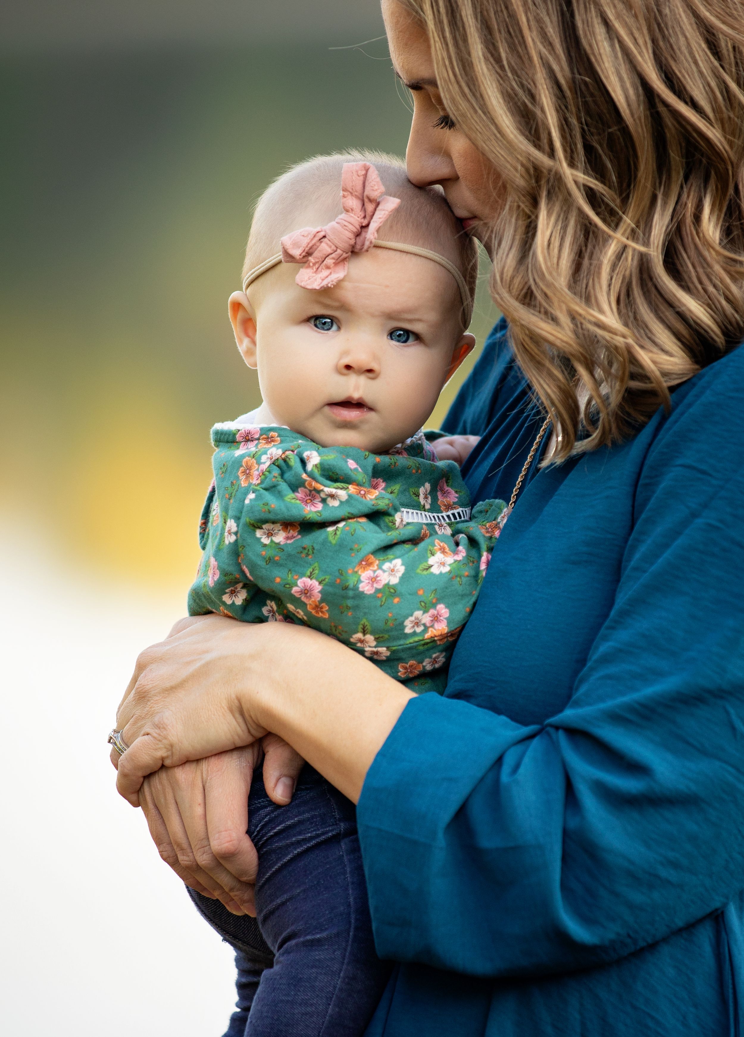 A mother in a teal top kisses her baby girl wearing a floral outfit and pink bow headband.