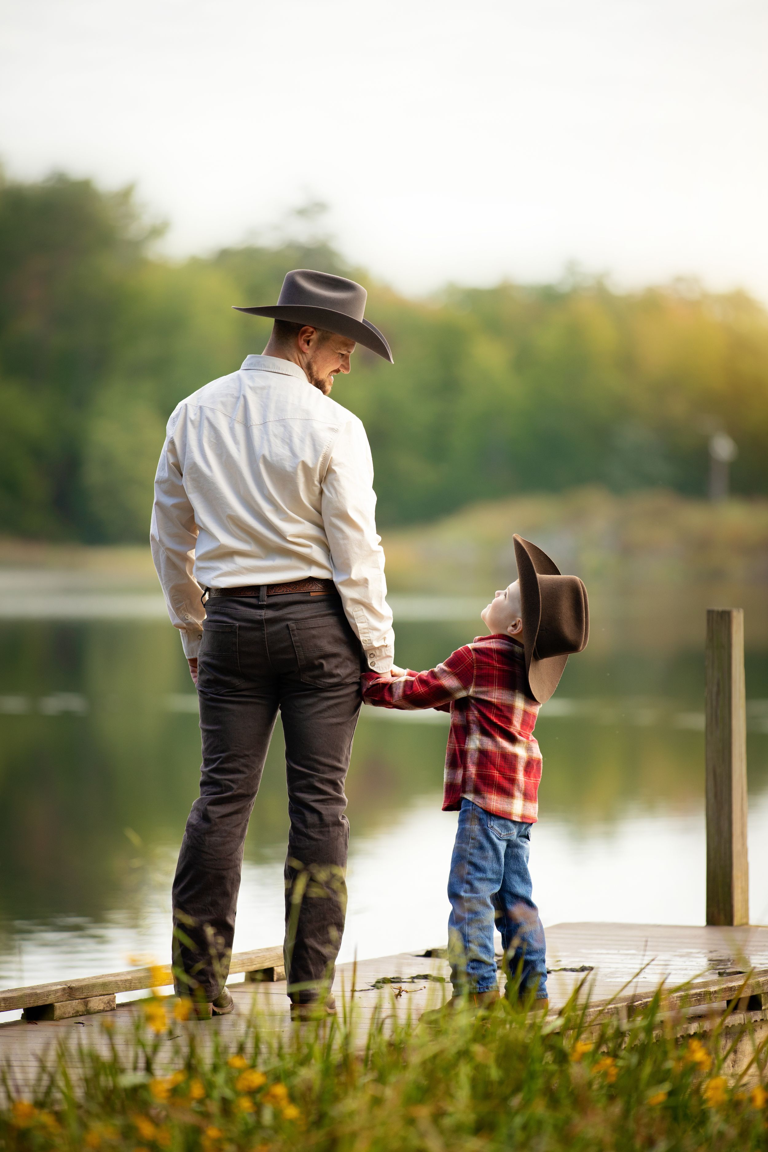 A father and young son in cowboy hats hold hands on a wooden dock overlooking a peaceful lake surrounded by trees.