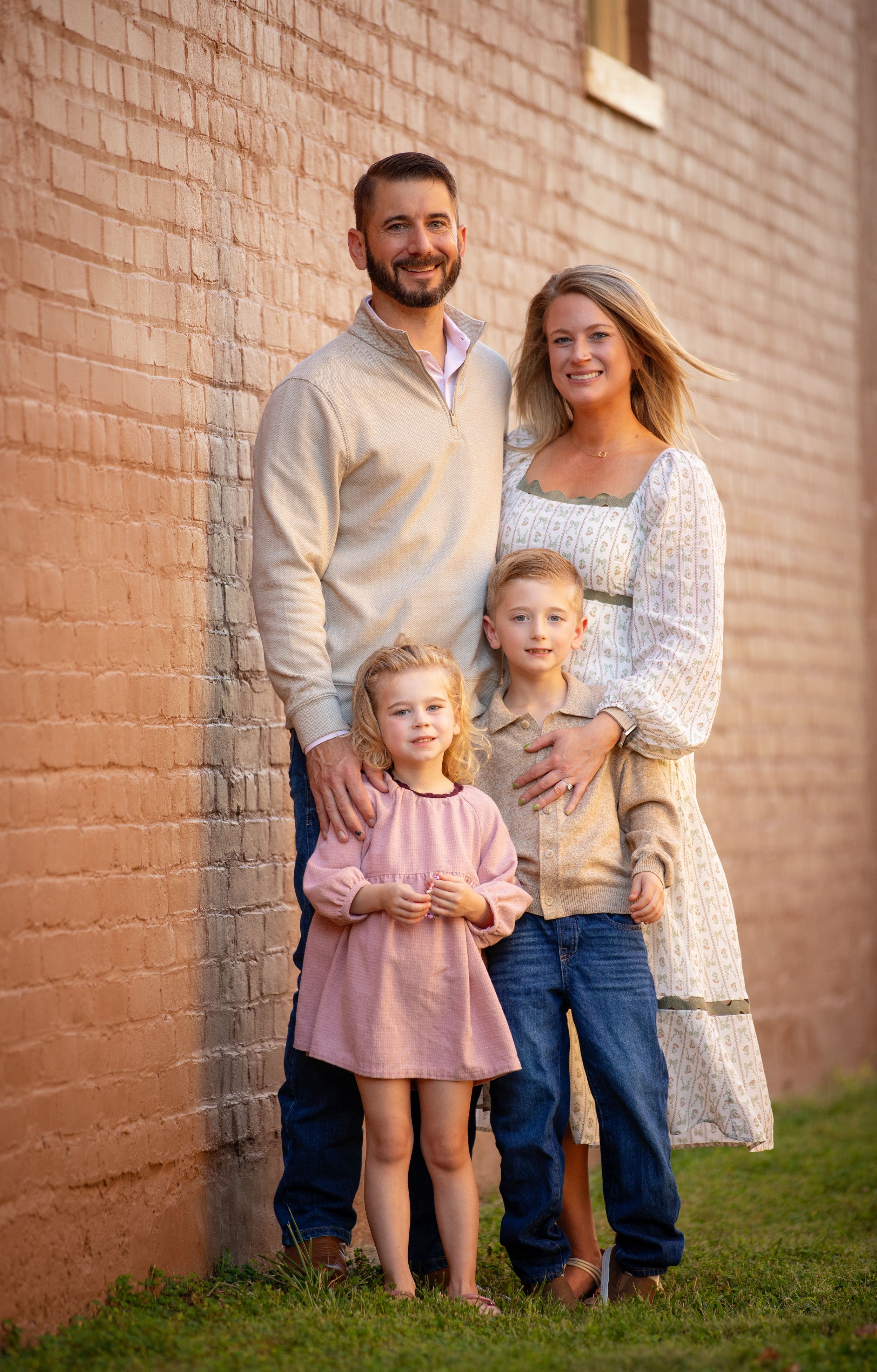 Family of four posing against a brick wall, parents with two young children smiling outdoors in casual attire.