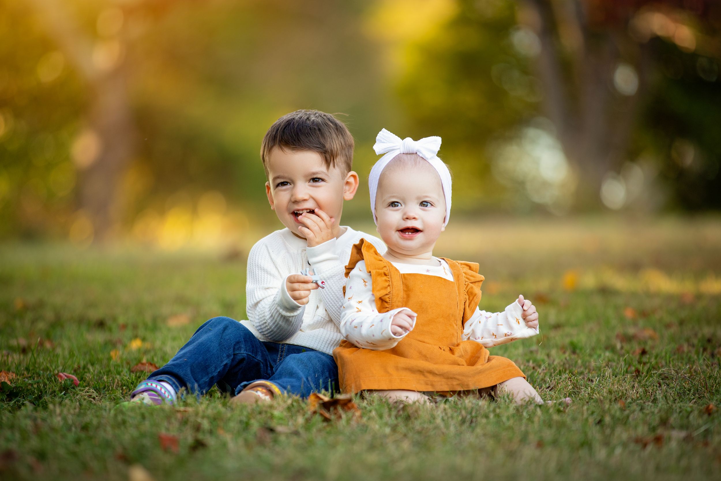 Two young children sit together in autumn grass surrounded by golden fall foliage.