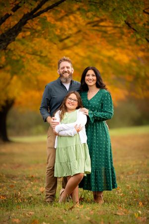 A family poses together on fallen autumn leaves with vibrant orange foliage in the background.