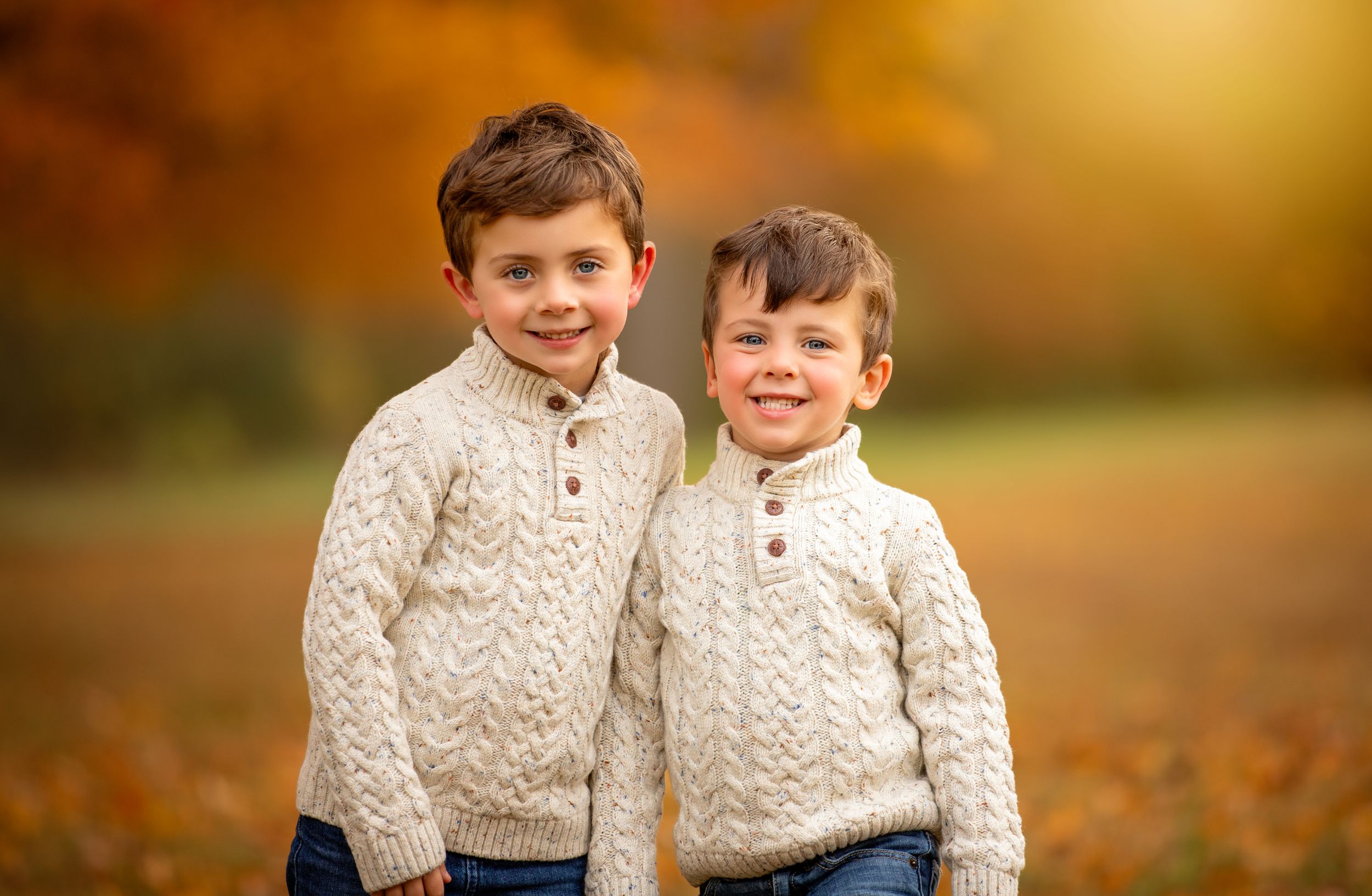 Children wearing matching cream cable-knit sweaters play in autumn leaves during golden hour.