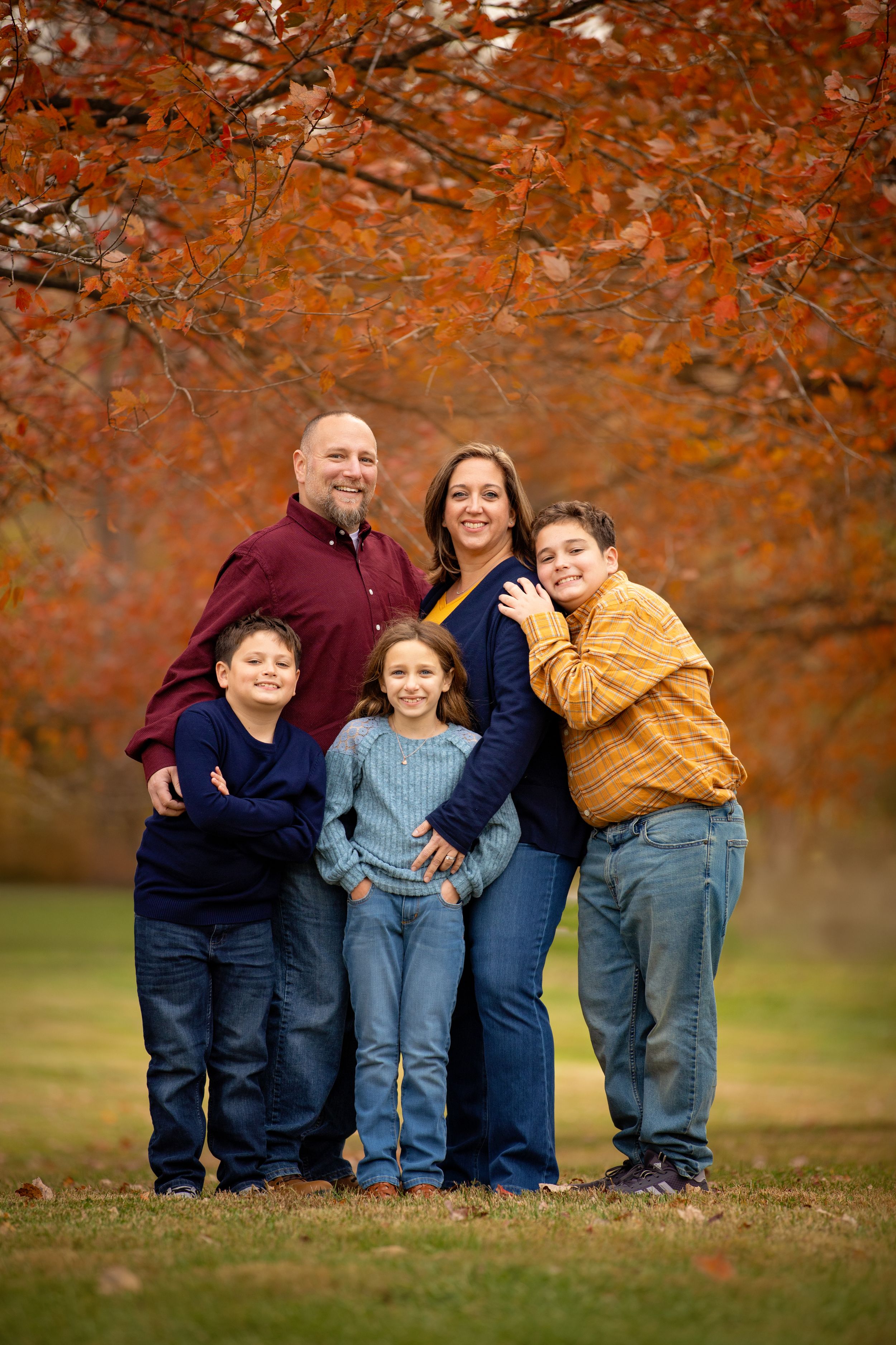 A family poses together under vibrant orange autumn foliage in a park setting.