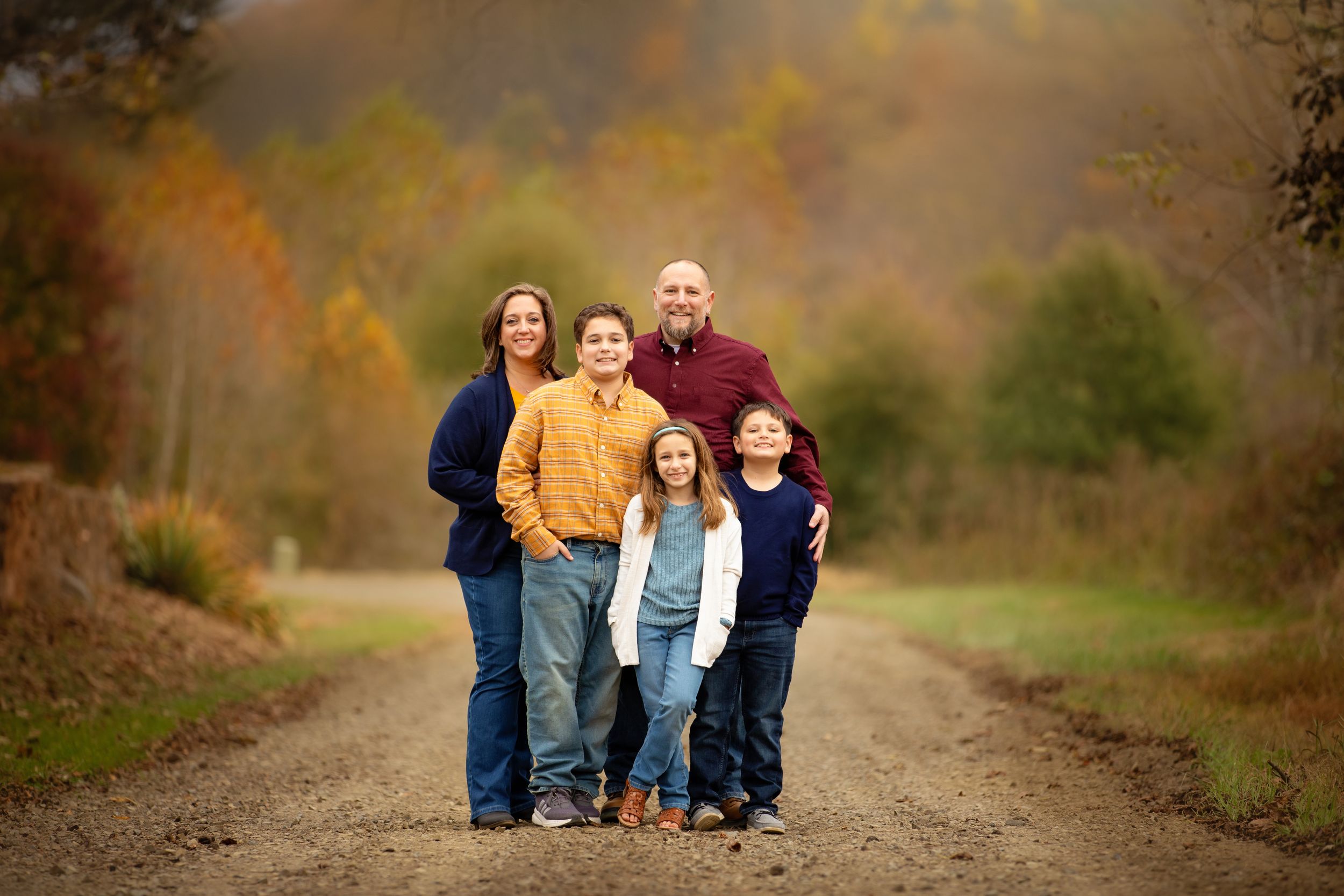 Family poses together on a dirt path surrounded by autumn foliage and warm fall colors.