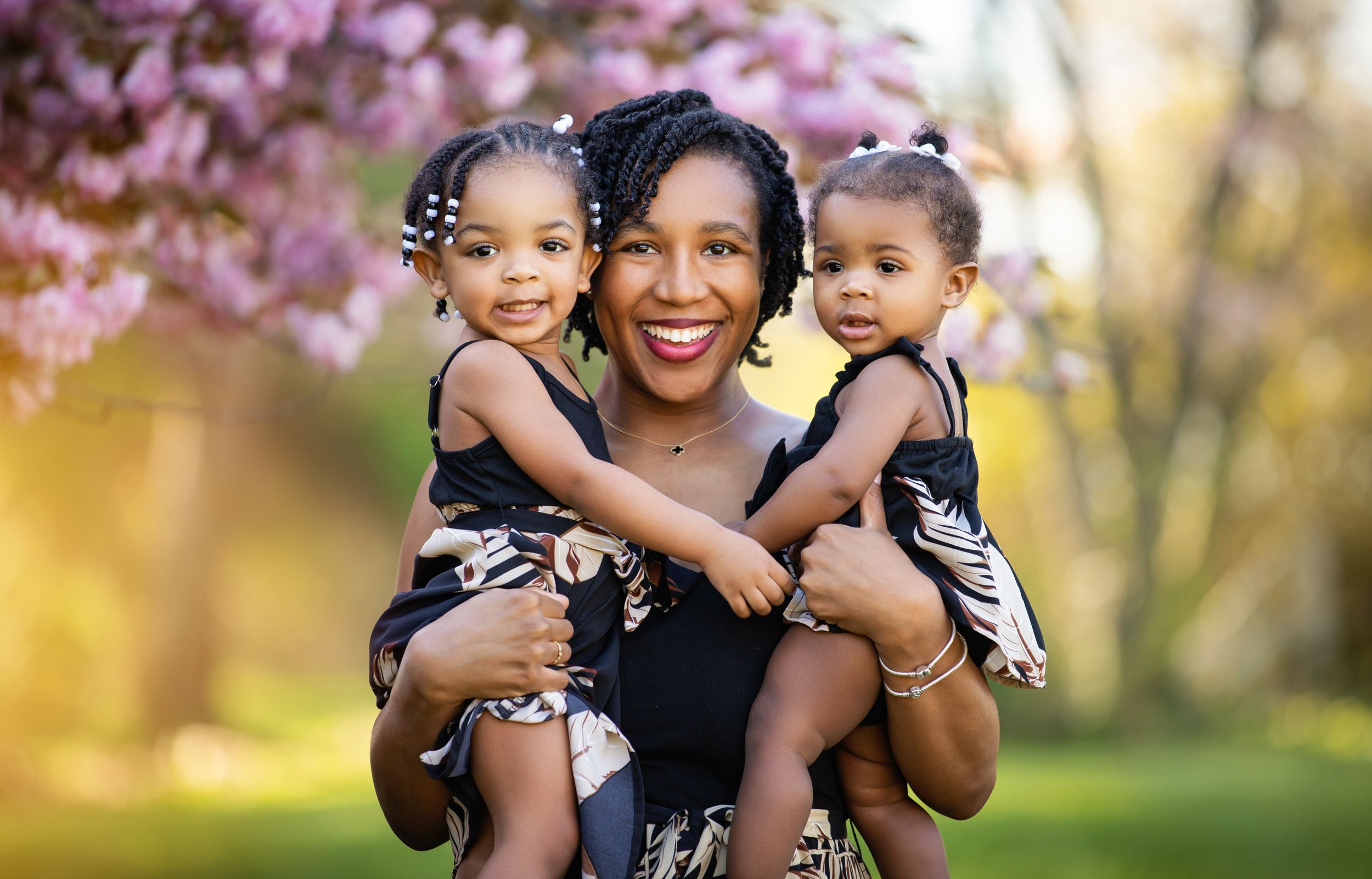 Three family members embrace and smile together in coordinated black outfits against a spring garden backdrop with pink blooms.