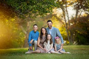 A family sits together on green grass in a park during golden hour sunset with trees in the background.