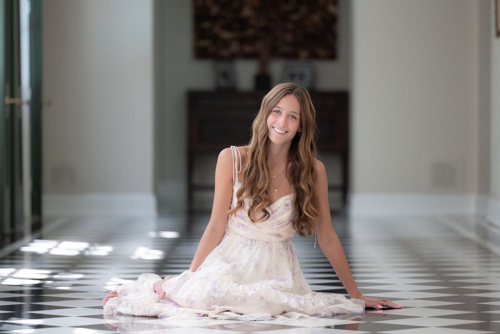 Young woman in white dress sitting gracefully on black and white checkered floor, smiling in elegant hallway.