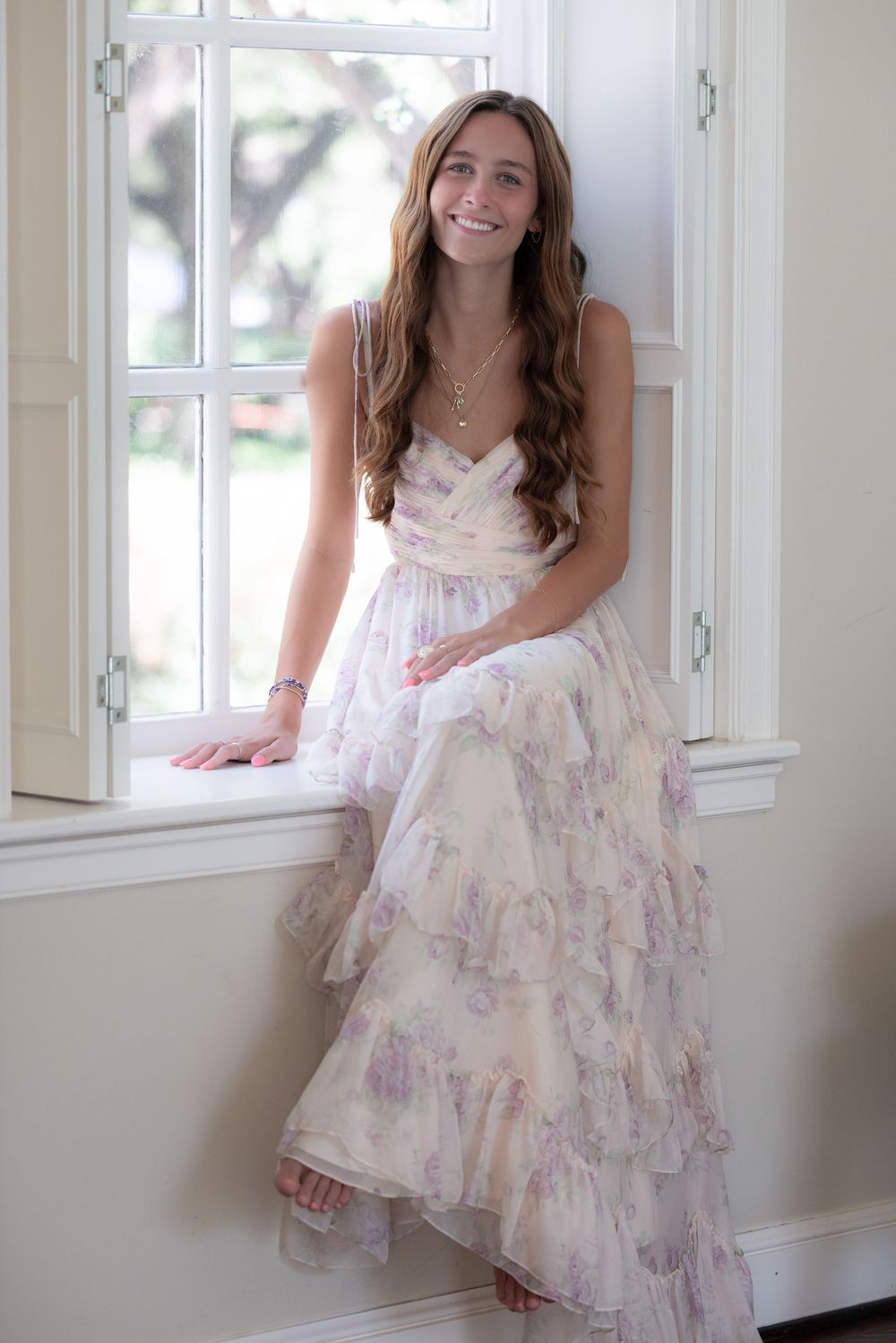 Young woman in a floral ruffled maxi dress smiling while sitting on a window sill in a bright room.