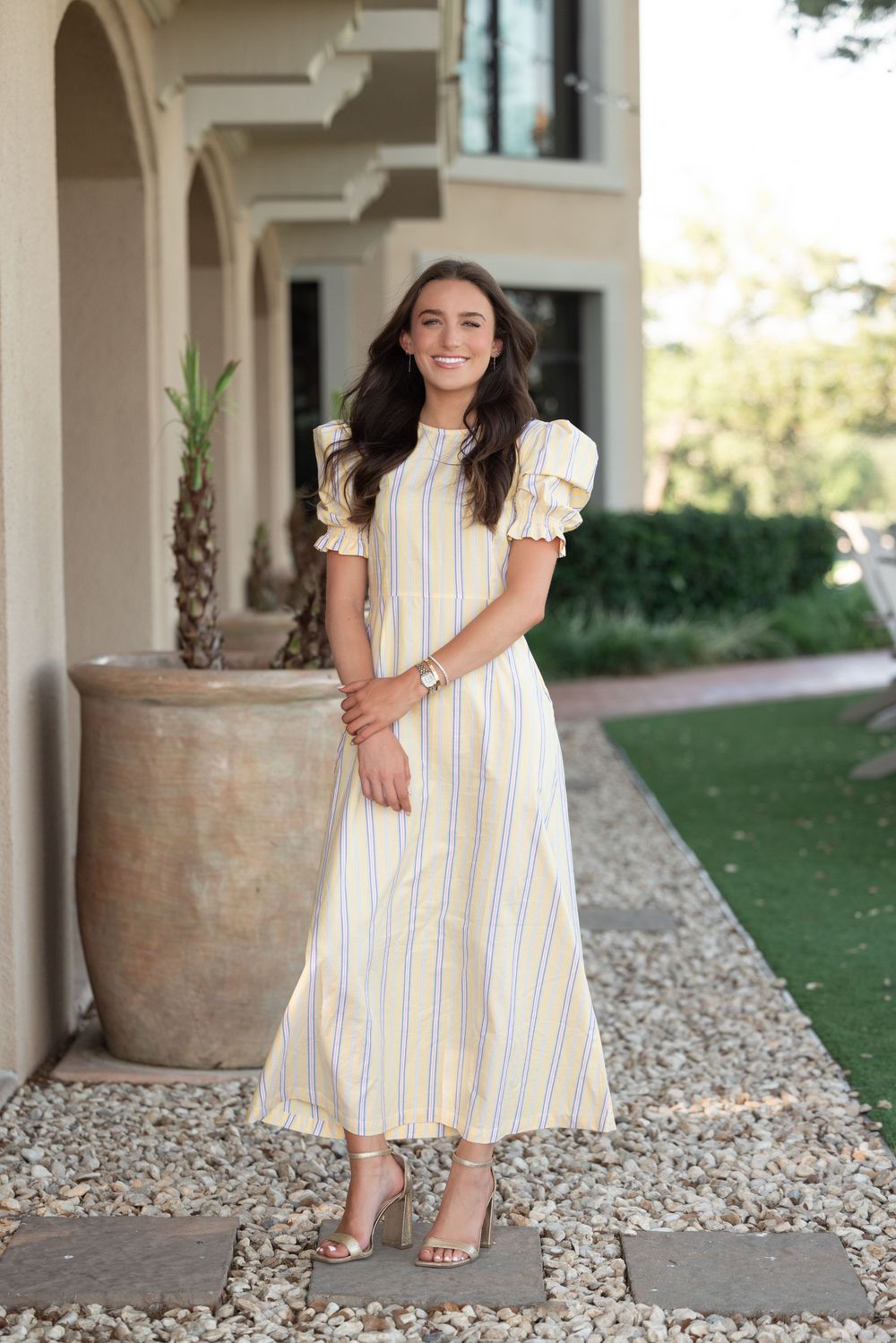 Woman in yellow striped midi dress with puff sleeves and heeled sandals standing on a gravel path outside a home.
