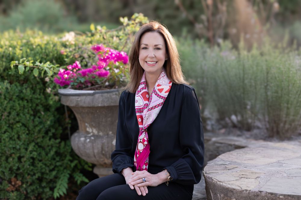 Professional woman in black blazer and floral scarf smiling outdoors in a garden setting with pink flowers.