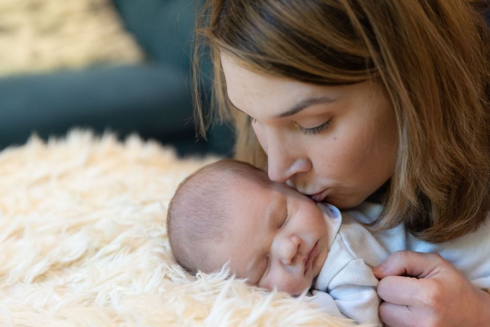 A tender moment as a young mother gently kisses her sleeping newborn baby resting on a soft fluffy white blanket.