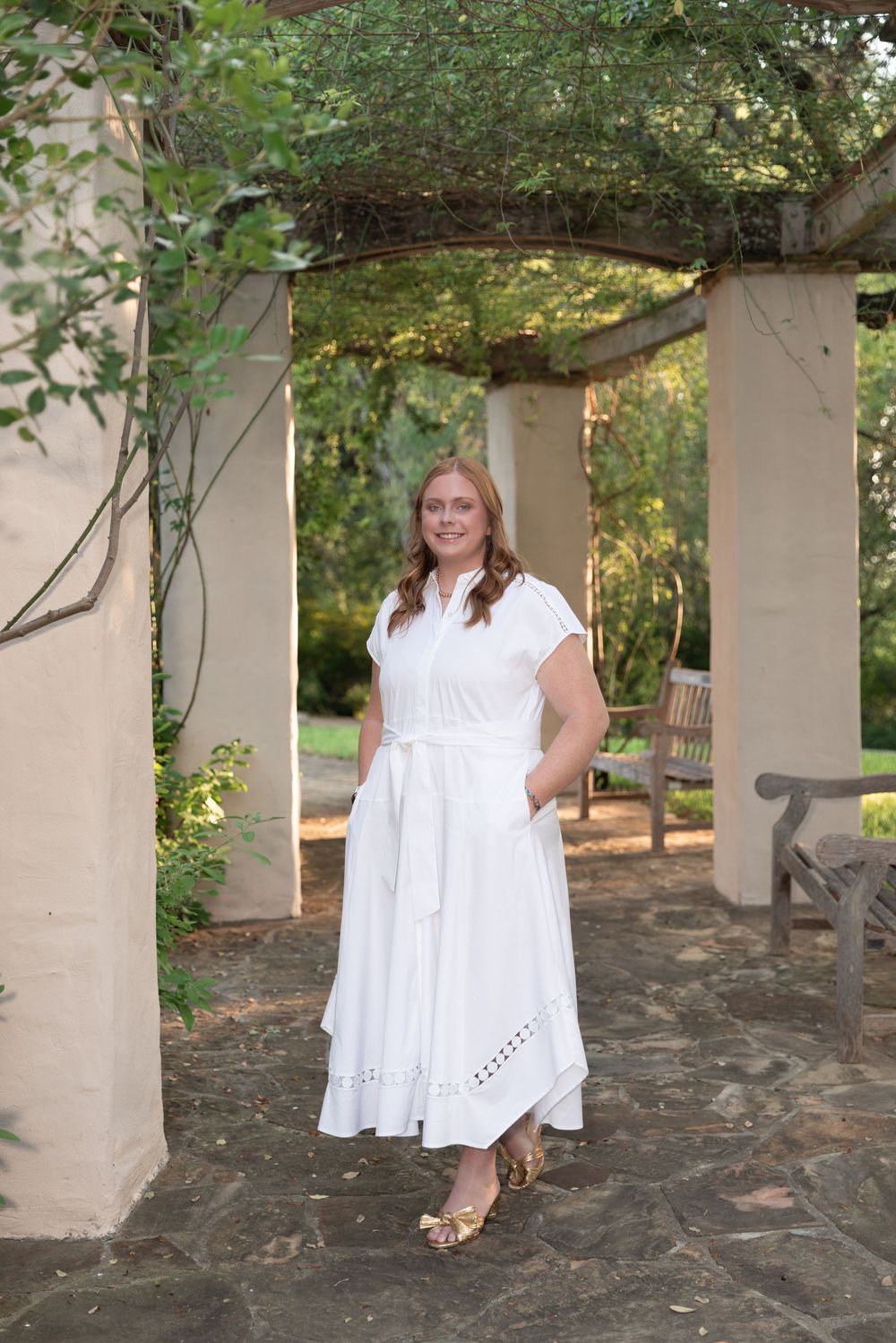 Woman in white belted midi dress standing under garden pergola with stone pillars and lush greenery.