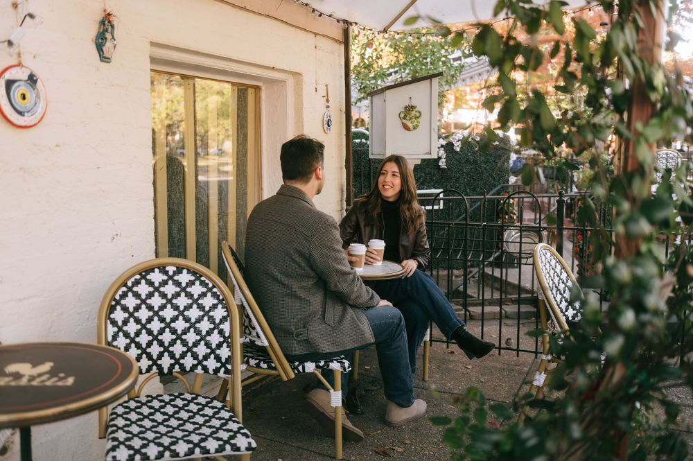 Couple enjoying coffee on a patio with black and white patterned chairs and greenery.