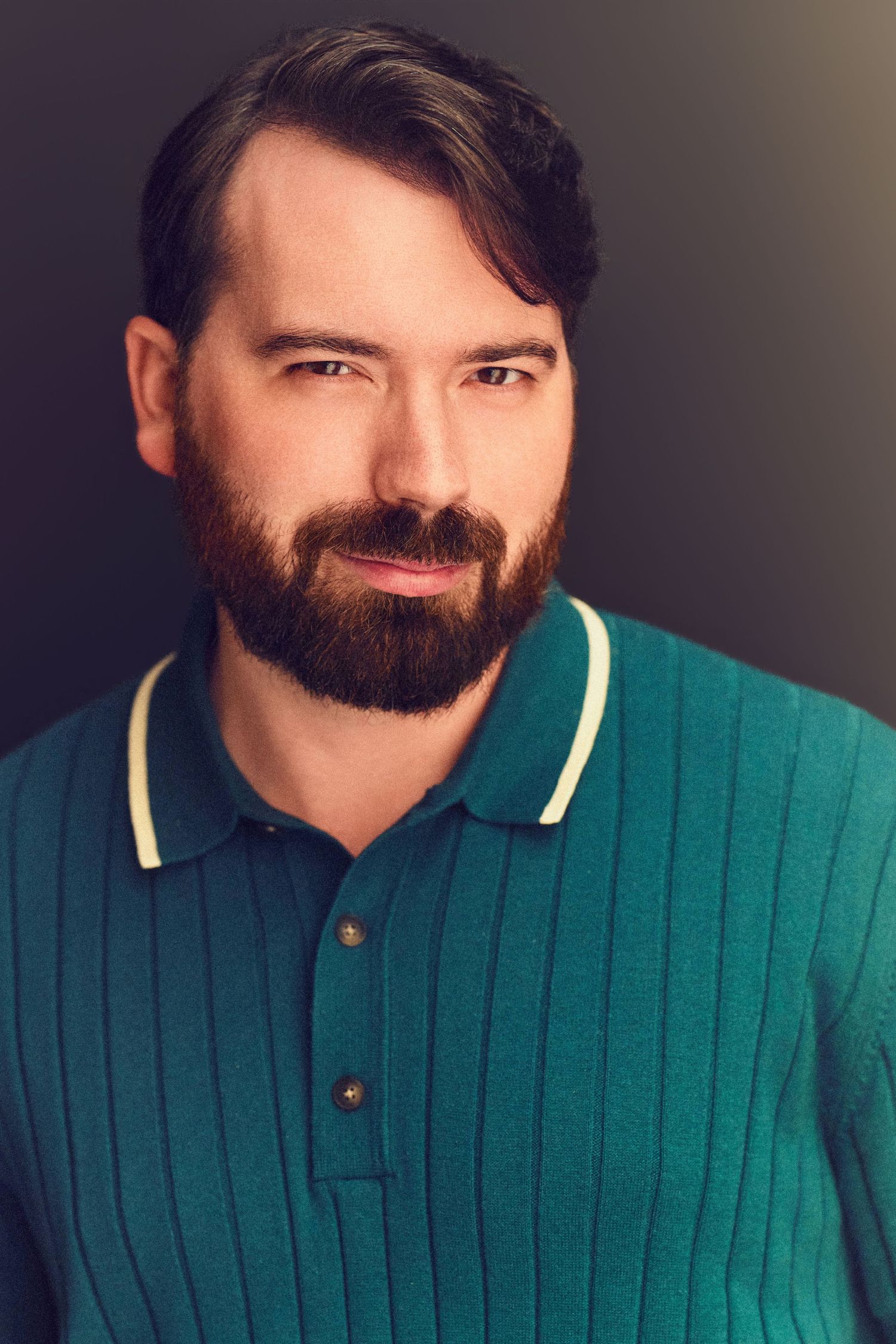 Professional headshot of a bearded man with dark hair wearing a teal ribbed polo shirt against a dark background.