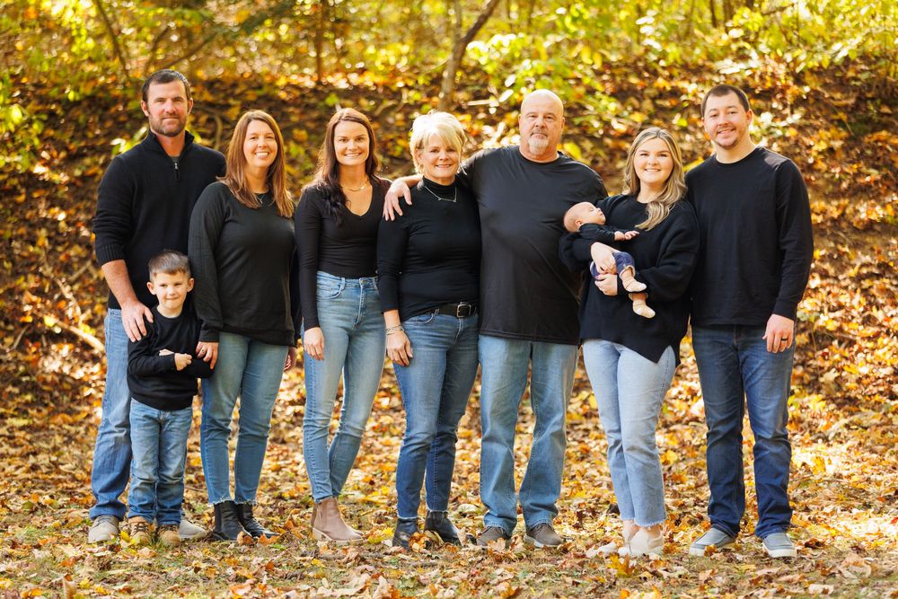 Multi-generational family portrait outdoors in autumn, eight members in matching black tops and jeans among fall foliage.