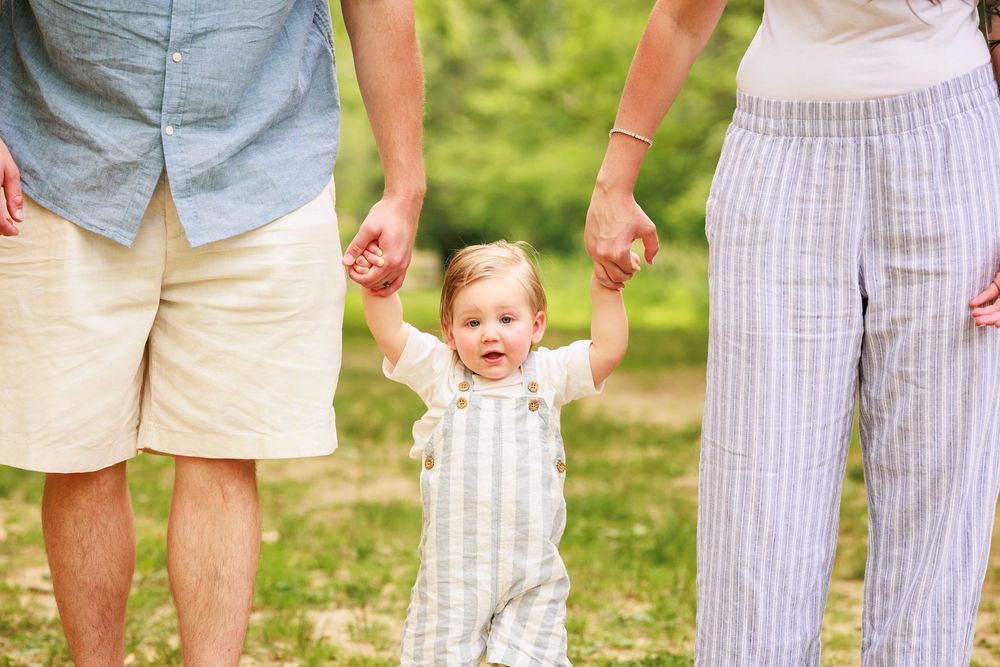 Two parents holding hands with a happy toddler in striped overalls learning to walk outdoors.