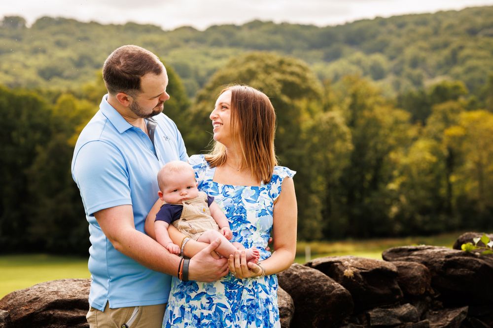 A young family poses outdoors, parents holding their newborn baby with lush green hills in the background.