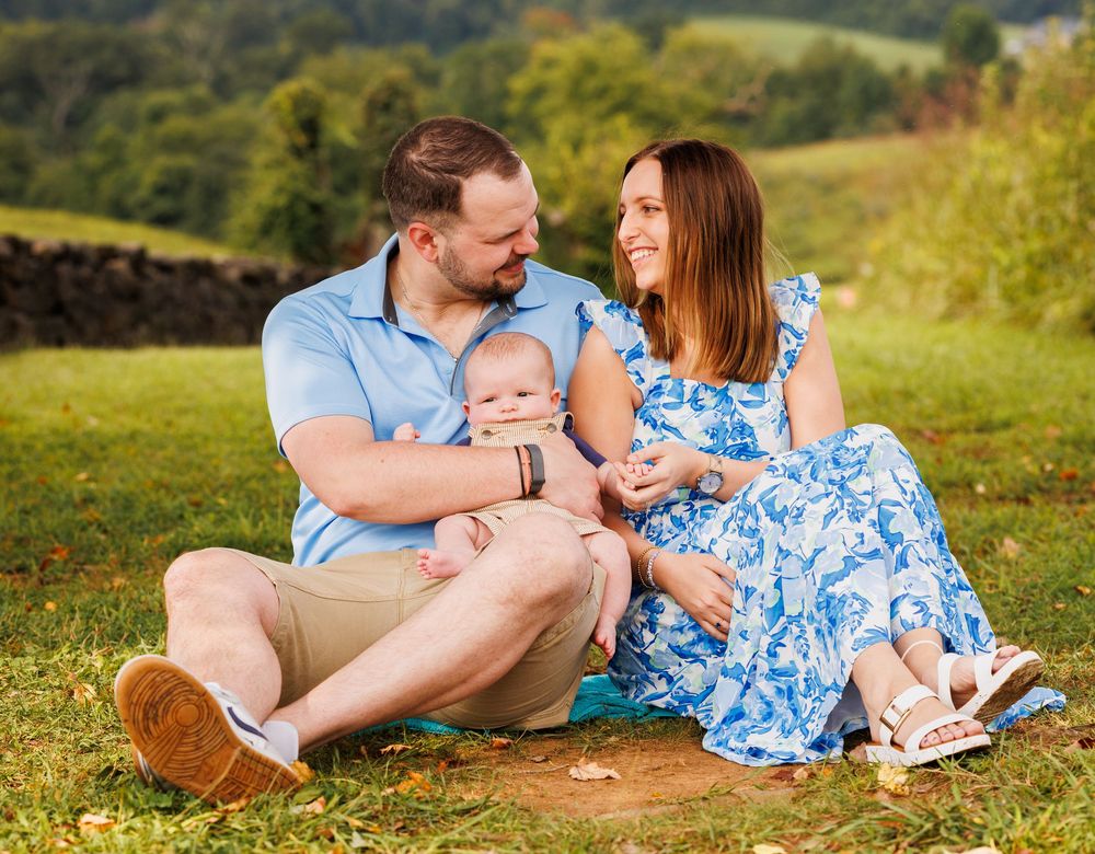 Happy family with parents and baby sitting on grass with scenic green hills in background.