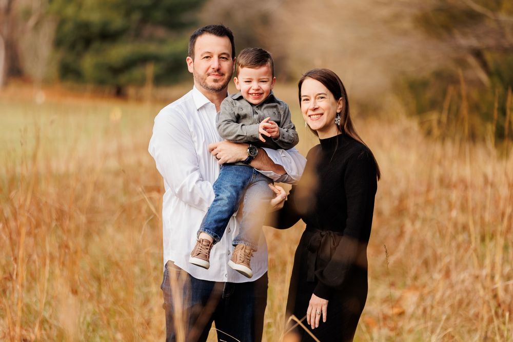 Family of three smiling outdoors in a golden field, parents holding toddler boy, autumn nature background.