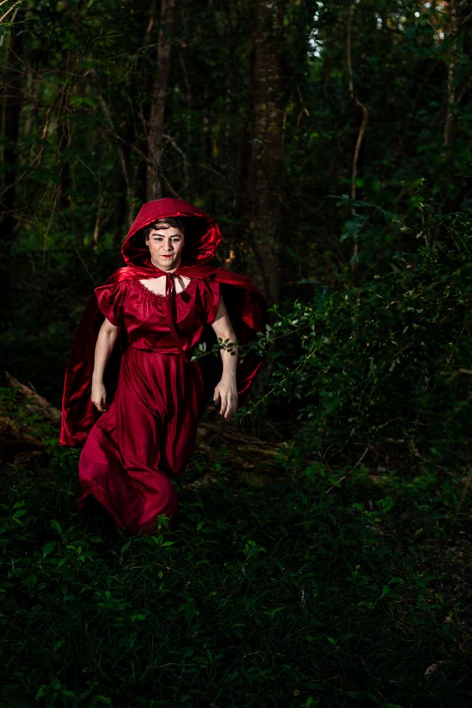 A figure in a flowing red dress and hood walks mysteriously through a dark forest setting.