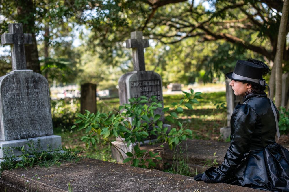 A person in Victorian-style black clothing sits pensively among old gravestones in a cemetery.