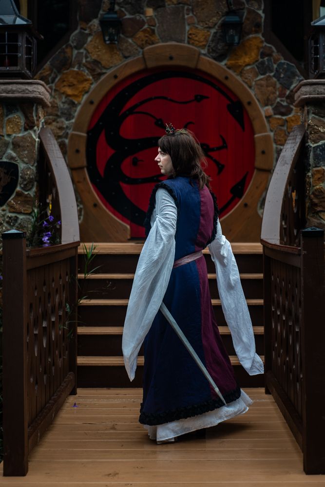 A figure in a flowing white dress and navy blue gown dances dramatically on a rustic wooden bridge with red archways behind.