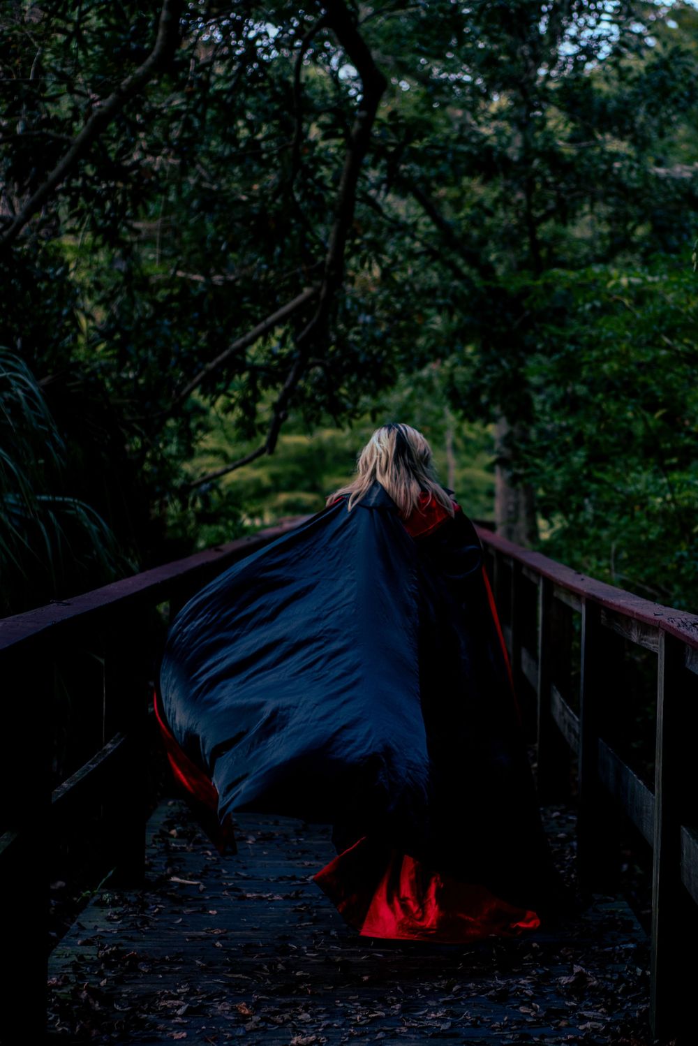 A flowing blue dress contrasts with red boots on a dark wooden footbridge surrounded by lush forest at dusk.