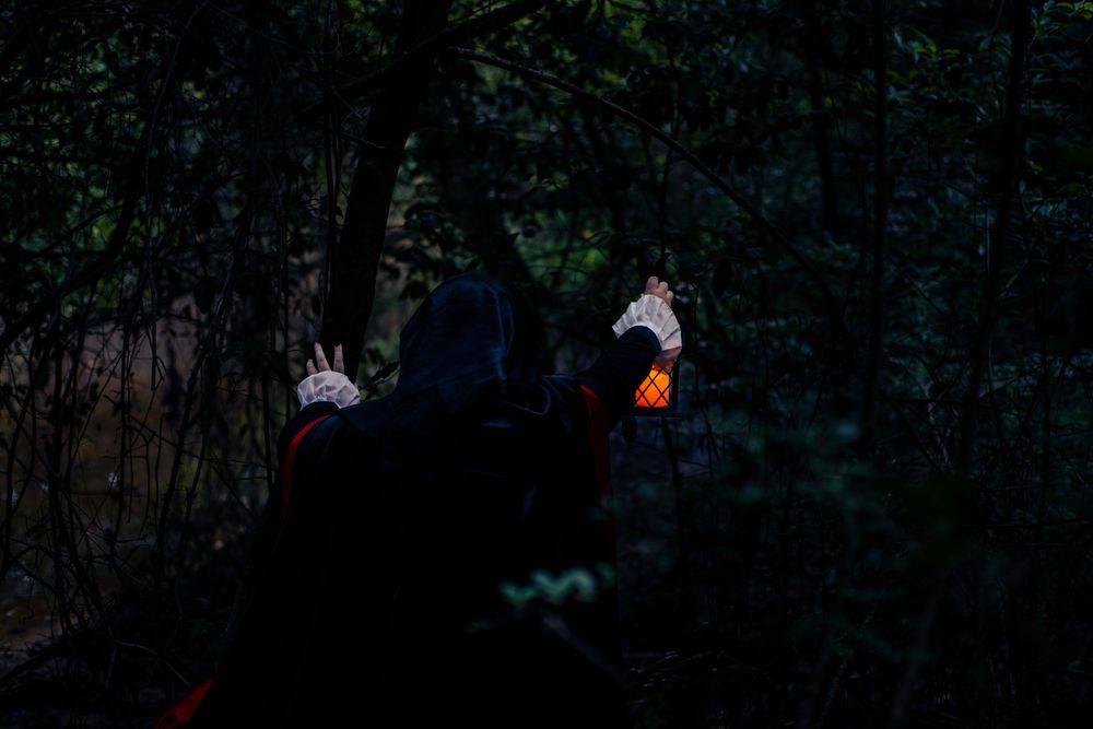 Dark mysterious figure in a hooded cloak holding glowing puppets stands in spooky forest at night.