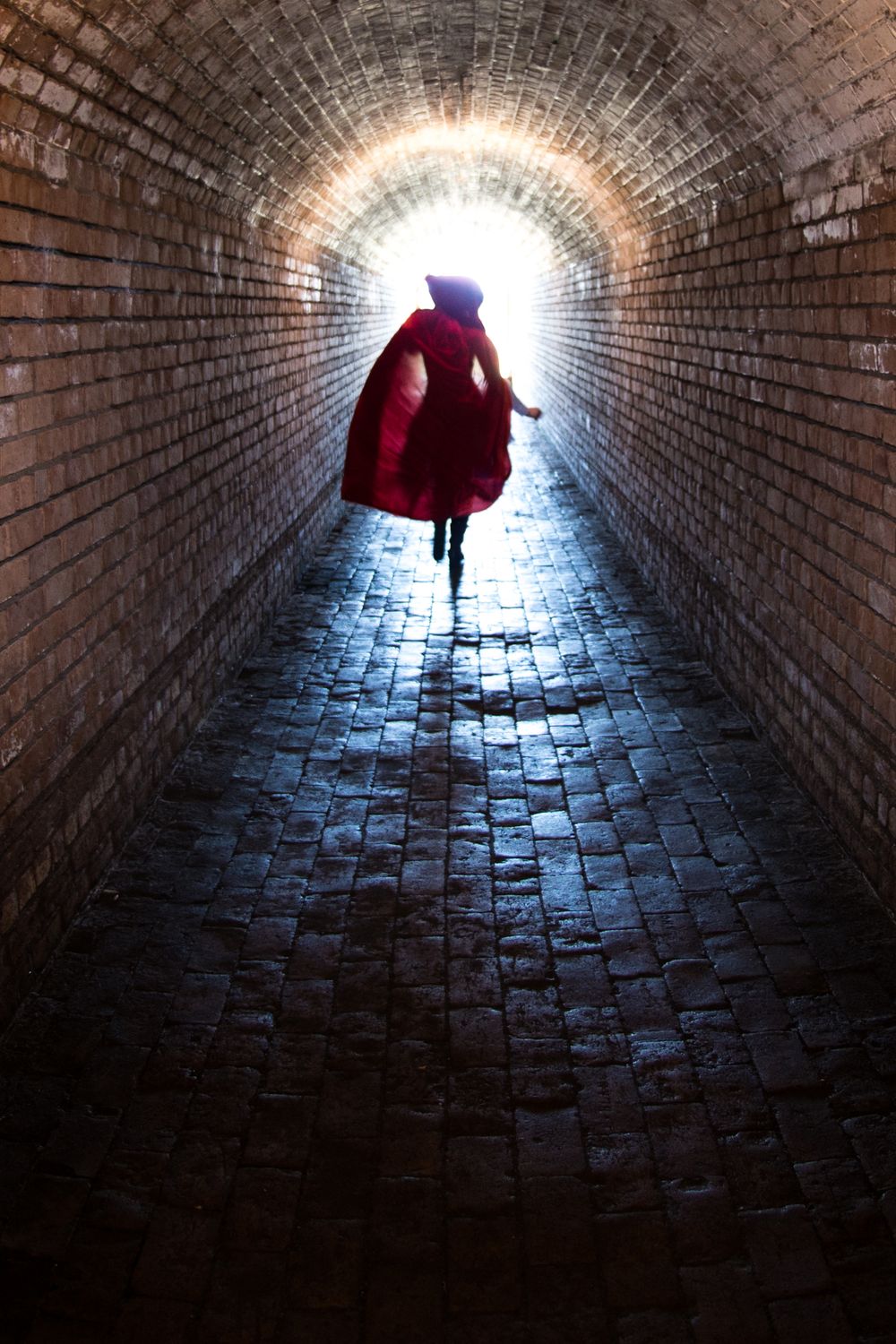 A silhouetted figure in a red coat walks through a dark brick tunnel toward a bright light at the end.