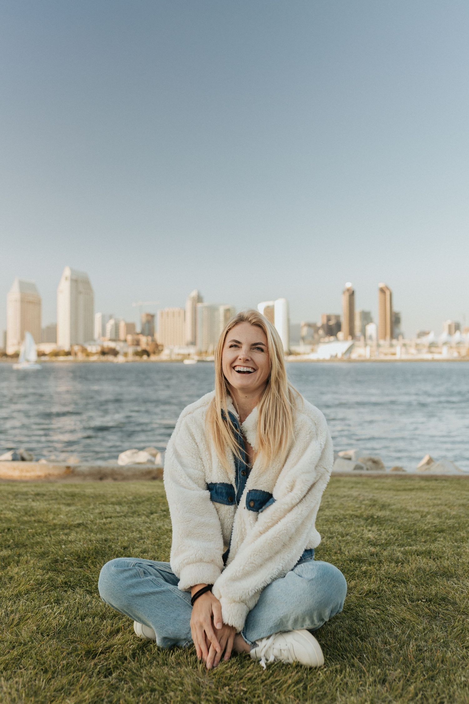 A woman sitting crossed legged on grass laughing with water and the San Diego cityscape.