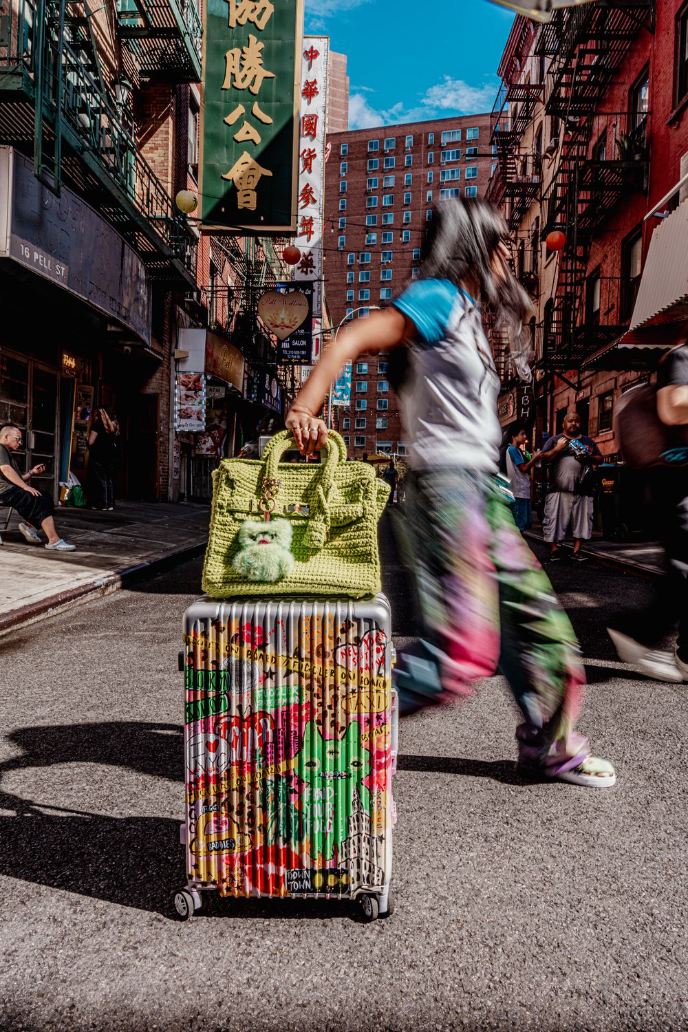 Person pulling colorful floral luggage with green bag through busy Chinatown street in NYC.
