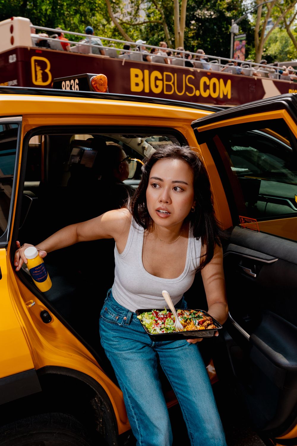 A young woman exits a yellow NYC taxi holding a salad bowl and drink with a Big Bus tour in the background.
