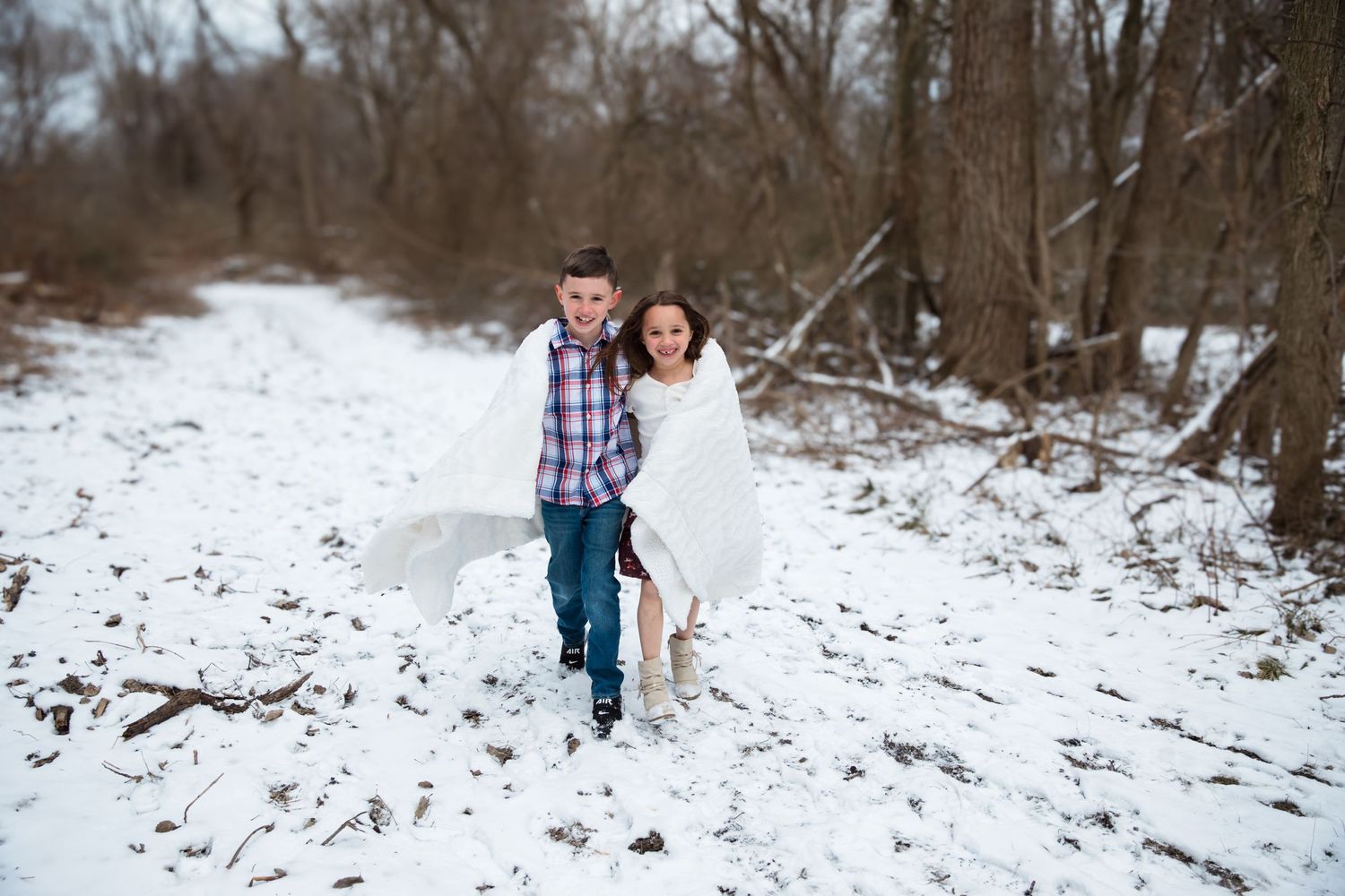 A couple poses together on a snowy winter path surrounded by bare trees.