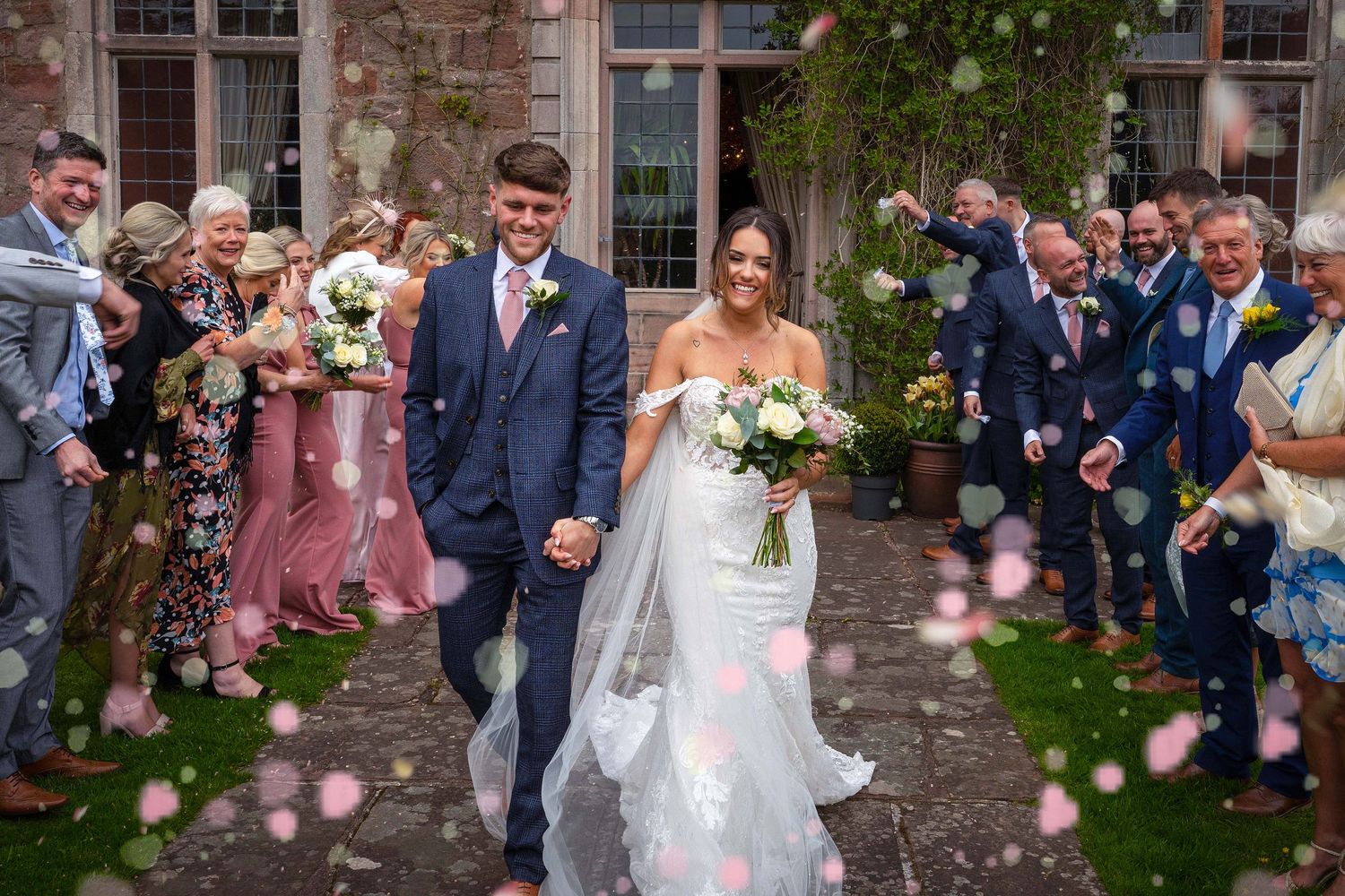 A couple walks down a petal-lined path surrounded by guests at an elegant English manor wedding celebration.