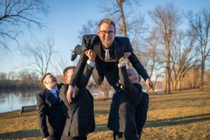 Wedding party lifts and carries person on their shoulders in outdoor park setting.