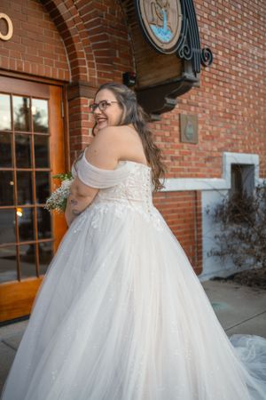 Bride in white ball gown smiles over shoulder against brick building backdrop.
